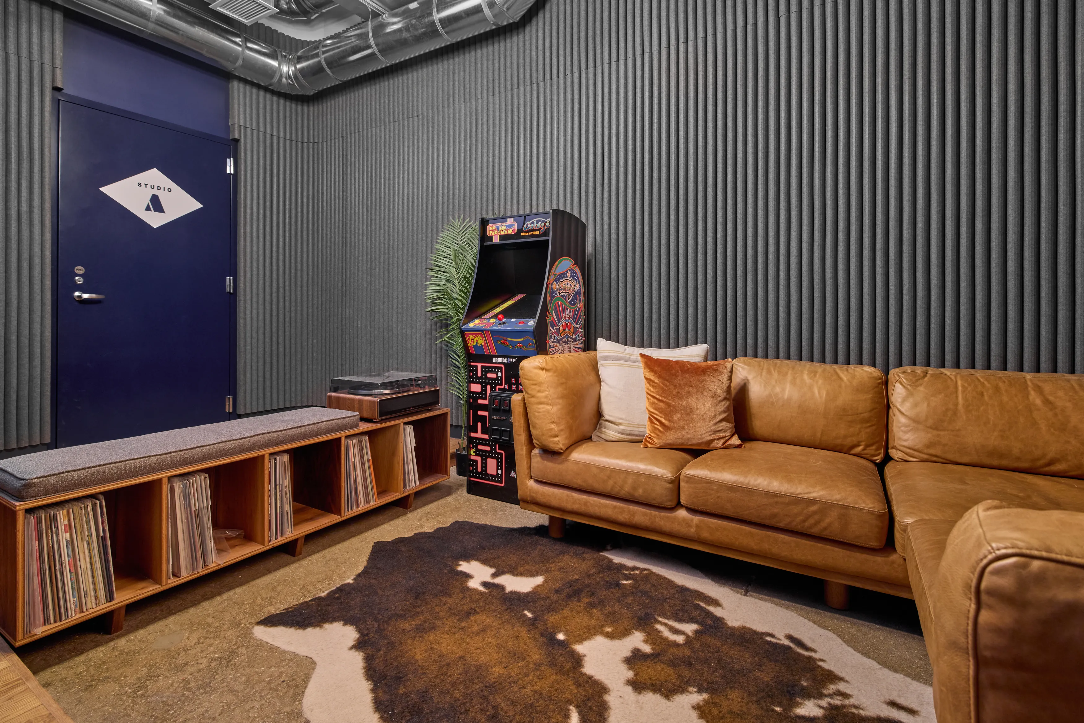 Wide shot showing a door to Studio A, a bench with vinyl records stored beneath, an arcade machine, and a L shaped couch.